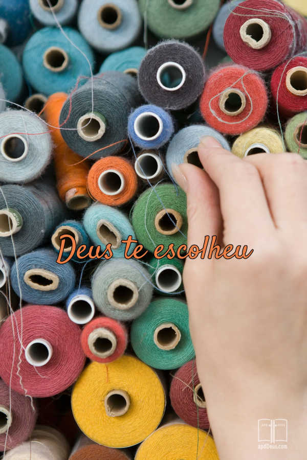 A fotografia mostra uma mão escolhendo entre bobinas de fios coloridos, dispostas de forma densa. No centro, um texto em laranja, escrito em letra cursiva, diz “Deus escolheu você”, enquanto fios soltos, tubetes de papelão e fibras variadas — nas cores vermelha, azul, verde, amarela, cinza e branca — compõem os detalhes visuais.