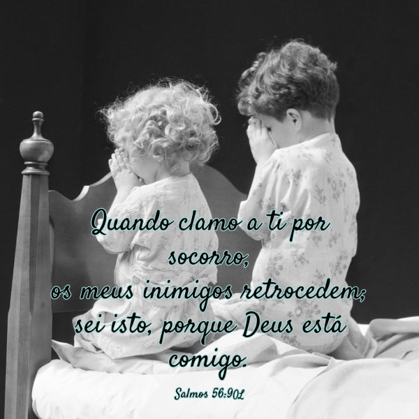 Duas crianças pequenas se ajoelham ao lado da cama para fazer orações antes de dormir. Salmos 56:9: "Meus inimigos recuarão quando eu clamar a ti por socorro. De uma coisa eu sei: Deus está do meu lado!"